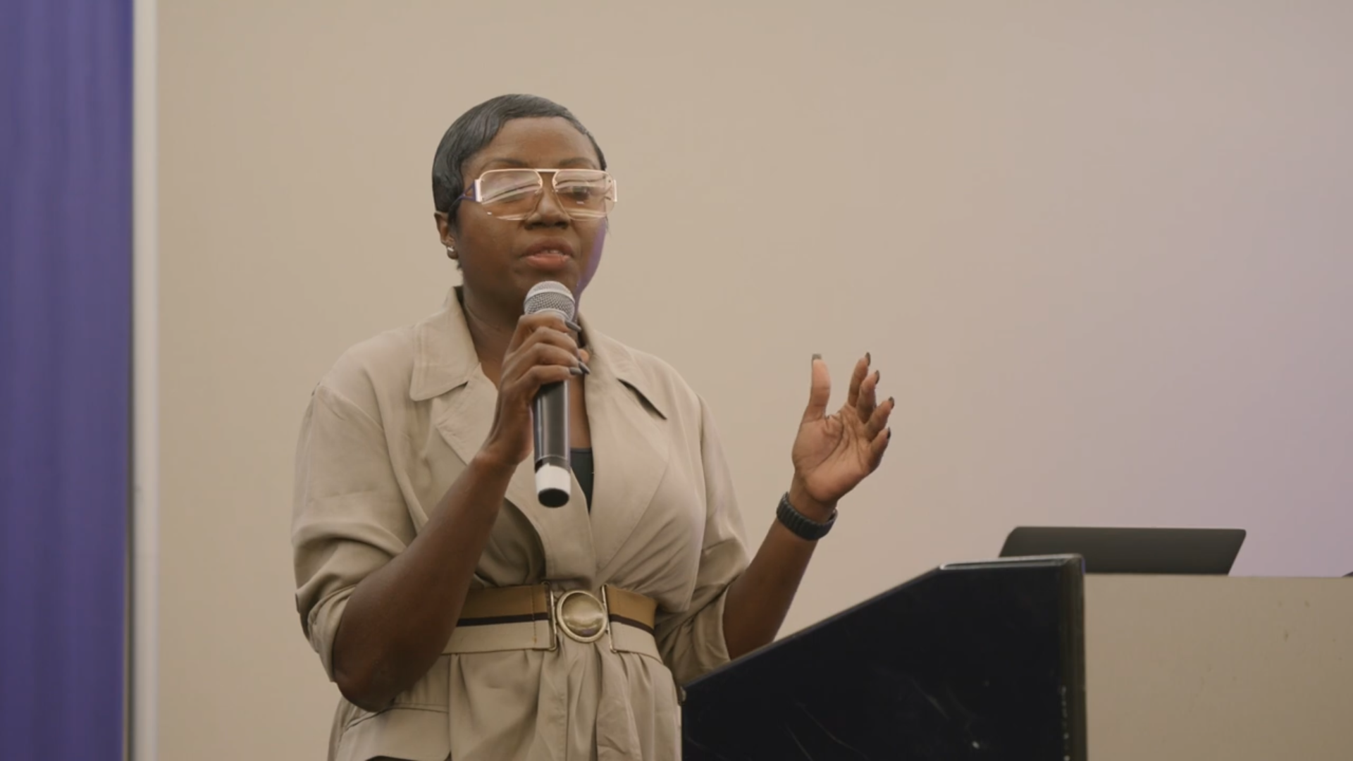 A person stands speaking into a microphone, wearing glasses and a beige outfit, gesturing with one hand at a podium.